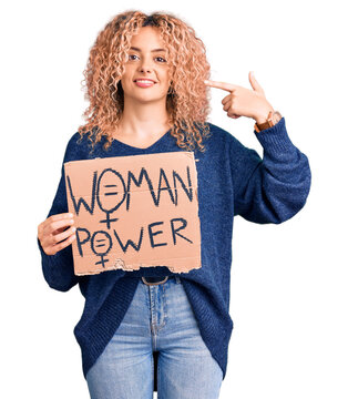 Young blonde woman with curly hair holding we need a change banner pointing finger to one self smiling happy and proud