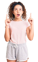 Beautiful kid girl with curly hair wearing casual clothes amazed and surprised looking up and pointing with fingers and raised arms.