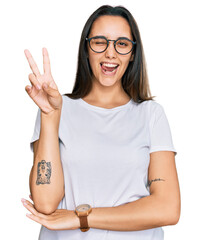 Young hispanic woman wearing casual white t shirt smiling with happy face winking at the camera doing victory sign with fingers. number two.
