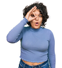 Young hispanic woman with curly hair wearing casual clothes doing ok gesture shocked with surprised face, eye looking through fingers. unbelieving expression.