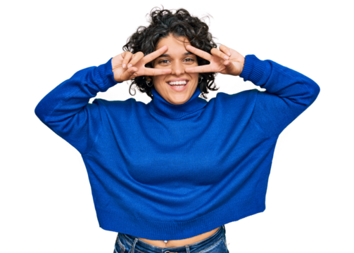 Young hispanic woman with curly hair wearing turtleneck sweater doing peace symbol with fingers over face, smiling cheerful showing victory