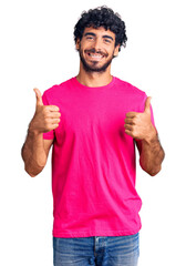 Handsome young man with curly hair and bear wearing casual pink tshirt success sign doing positive gesture with hand, thumbs up smiling and happy. cheerful expression and winner gesture.