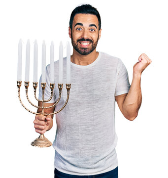 Young hispanic man with beard holding menorah hanukkah jewish candle screaming proud, celebrating victory and success very excited with raised arm