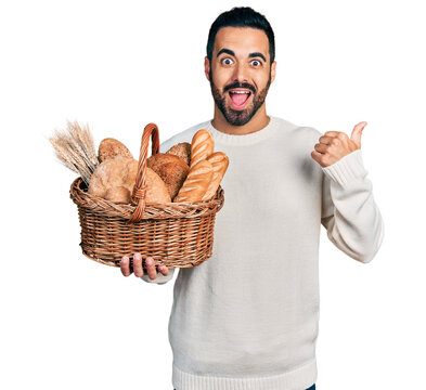 Young hispanic man with beard holding wicker basket with bread pointing thumb up to the side smiling happy with open mouth
