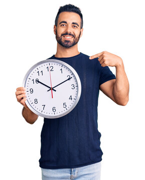 Young hispanic man holding big clock pointing finger to one self smiling happy and proud