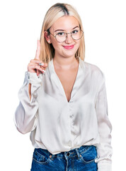 Beautiful blonde woman wearing elegant shirt and glasses showing and pointing up with finger number one while smiling confident and happy.