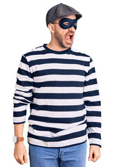 Young handsome man wearing burglar mask angry and mad screaming frustrated and furious, shouting with anger. rage and aggressive concept.