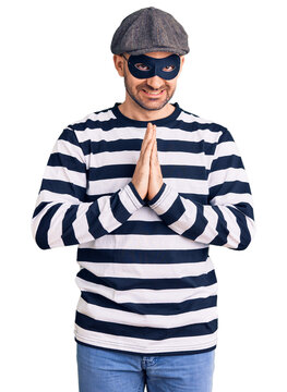 Young handsome man wearing burglar mask praying with hands together asking for forgiveness smiling confident.