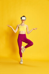 Fototapeta na wymiar Full-length image of young slim woman wearing vr glases and training against yellow studio background. Yoga and meditation. Concept of sport, fitness, body care, fashion, youth, lifestyle, ad