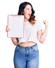 Beautiful young brunette woman holding clipboard with blank space pointing thumb up to the side smiling happy with open mouth