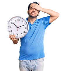 Young hispanic man holding big clock stressed and frustrated with hand on head, surprised and angry...