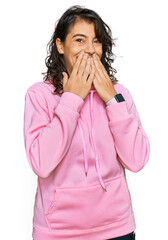Young hispanic woman wearing casual sweatshirt laughing and embarrassed giggle covering mouth with...