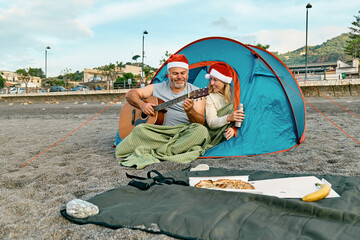 Middle aged couple wearing santa hats having christmas picnic on winter beach. Bearded man and pretty blond woman sitting in tent, playing guitar, talking and eating pizza.