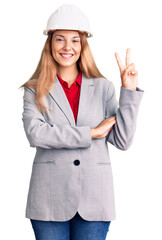 Beautiful young woman wearing architect hardhat smiling with happy face winking at the camera doing victory sign. number two.