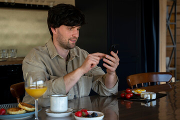 Smiling man sitting at a table in the kitchen with a cup of coffee and looking at a smartphone. A man with glasses enjoys breakfast. 