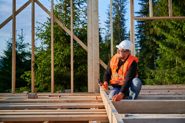 Wooden frame abode is being built by carpenter. The bearded man wearing glasses is driving nails...