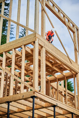 Wooden two-story frame house being built by carpenter. Man wearing glasses hammering nails using hammer, dressed in protective helmet and construction vest. Concept of modern ecological construction.