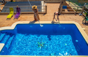 Two female friends and lesbian couple cleaning a swimming pool together dressed in sundress and bikini with a basket, brush and robotic pool vacuum cleaner on a sunny summer day