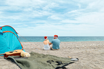 Back view of middle aged couple wearing santa hats having christmas picnic on the beach.