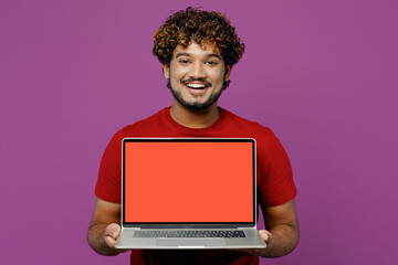 Young smiling happy IT Indian man he wear red t-shirt casual clothes hold use work on laptop pc computer with blank screen workspace area isolated on plain purple background studio. Lifestyle concept.