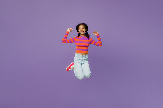 Full body happy overjoyed little kid teen girl 15-16 years old wear striped orange sweatshirt jump high do winner gesture clench fsit isolated on plain purple background. Childhood lifestyle concept.