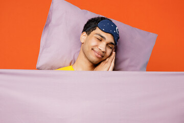 Calm young serene man wear pyjamas jam sleep eye mask rest relax at home lies wrap covered under blanket duvet close eyes isolated on plain orange color background studio. Good mood night nap concept.