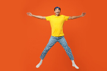 Fototapeta na wymiar Full body excited young man wear pyjamas jam sleep eye mask rest relax at home jump high with outstretched hands arms isolated on plain orange background studio portrait. Good mood night nap concept.