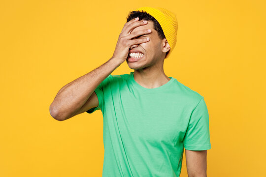 Young sad man of African American ethnicity he wearing casual clothes green t-shirt hat puts hand on face facepalm epic fail mistaken omg gesture isolated on plain yellow background studio portrait.