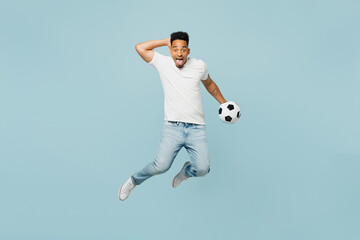 Obraz premium Full body shocked overjoyed happy fun young man fan wears t-shirt cheer up support football sport team hold soccer ball jump high watch tv live stream isolated on plain pastel blue color background.