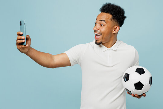 Young man fan wear basic t-shirt do selfie shot on mobile cell phone cheer up support football sport team hold in hand soccer ball watch tv live stream isolated on plain pastel blue color background.