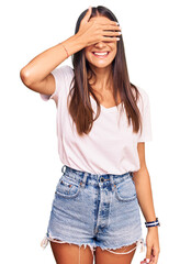 Young hispanic woman wearing casual white tshirt smiling and laughing with hand on face covering eyes for surprise. blind concept.