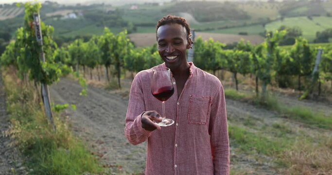 Happy young african man drinking red wine outdoor with vineyards in background - Tourist person smiling on camera during summer vacation - Travel and vine tasting concept