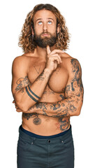 Fototapeta premium Handsome man with beard and long hair standing shirtless showing tattoos thinking concentrated about doubt with finger on chin and looking up wondering