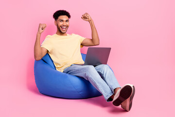 Full size photo of satisfied man wear yellow t-shirt jeans sit on bean bag with laptop win gambling...