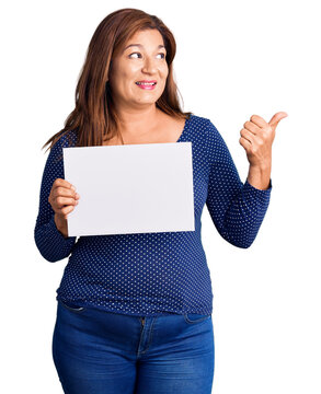 Middle age latin woman holding paper banner with blank space pointing thumb up to the side smiling happy with open mouth