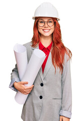 Young redhead architect woman wearing safety helmet holding blueprints looking positive and happy standing and smiling with a confident smile showing teeth