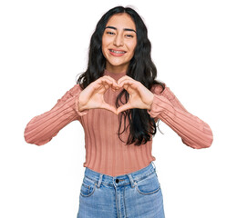 Hispanic teenager girl with dental braces wearing casual clothes smiling in love doing heart symbol...