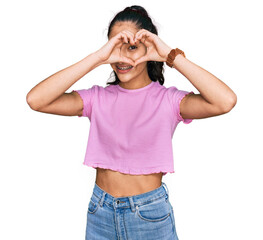 Hispanic teenager girl with dental braces wearing casual clothes doing heart shape with hand and fingers smiling looking through sign