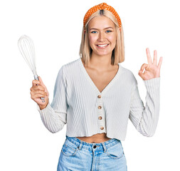 Beautiful young blonde woman holding baker whisk doing ok sign with fingers, smiling friendly gesturing excellent symbol