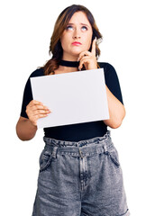 Young beautiful caucasian woman holding blank empty paper serious face thinking about question with hand on chin, thoughtful about confusing idea