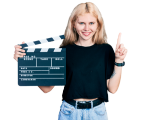 Young caucasian woman holding video film clapboard smiling with an idea or question pointing finger with happy face, number one