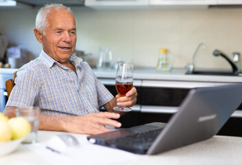 Happy elderly man with glass of wine at laptop sitting at table