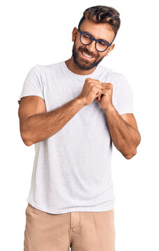 Young hispanic man wearing casual clothes and glasses laughing nervous and excited with hands on chin looking to the side