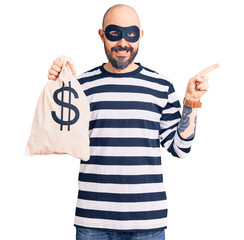 Young handsome man wearing burglar mask holding money bag with dollar symbol smiling happy pointing...