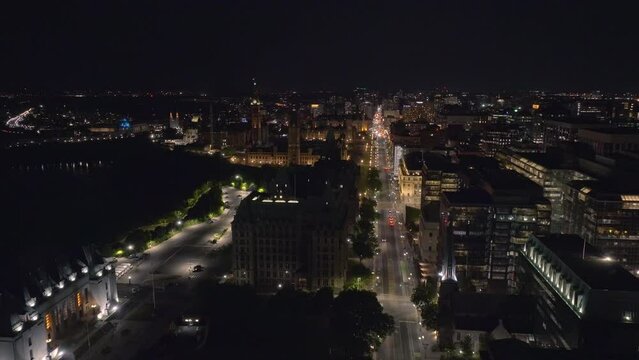 Slow backward aerial dolly shot of parliament buildings in downtown Ottawa with traffic driving on wellington street and city lights, the peace tower and parliament buildings are well lit.