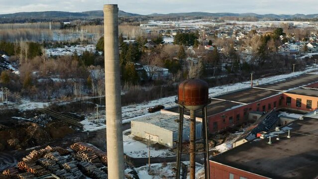 Abandoned industrial buildings in small town of Thurso in Quebec Canada. Old smokestack and rusted water tower as seen by a drone in a snow covered landscape.