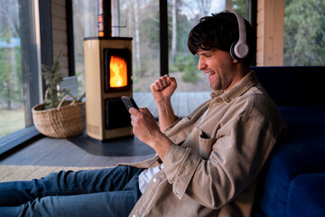 Young man is sitting on the floor by the sofa with headphones and a phone in his hands against the background of the fireplace. Listens to music, podcast. 