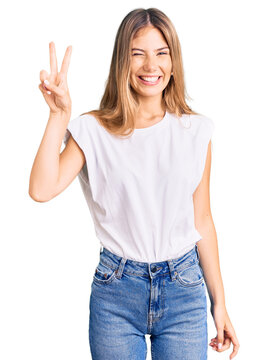 Beautiful caucasian woman with blonde hair wearing casual white tshirt smiling with happy face winking at the camera doing victory sign. number two.