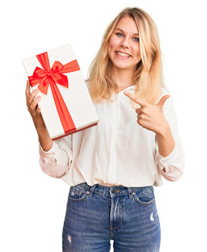 Young beautiful blonde woman holding gift pointing finger to one self smiling happy and proud