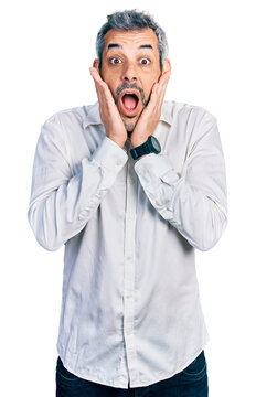 Middle age hispanic with grey hair wearing casual white shirt afraid and shocked, surprise and amazed expression with hands on face